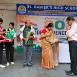 Group of students and adults on a school stage during a ceremony exchanging items with a banner reading st Xaviers high school in the background